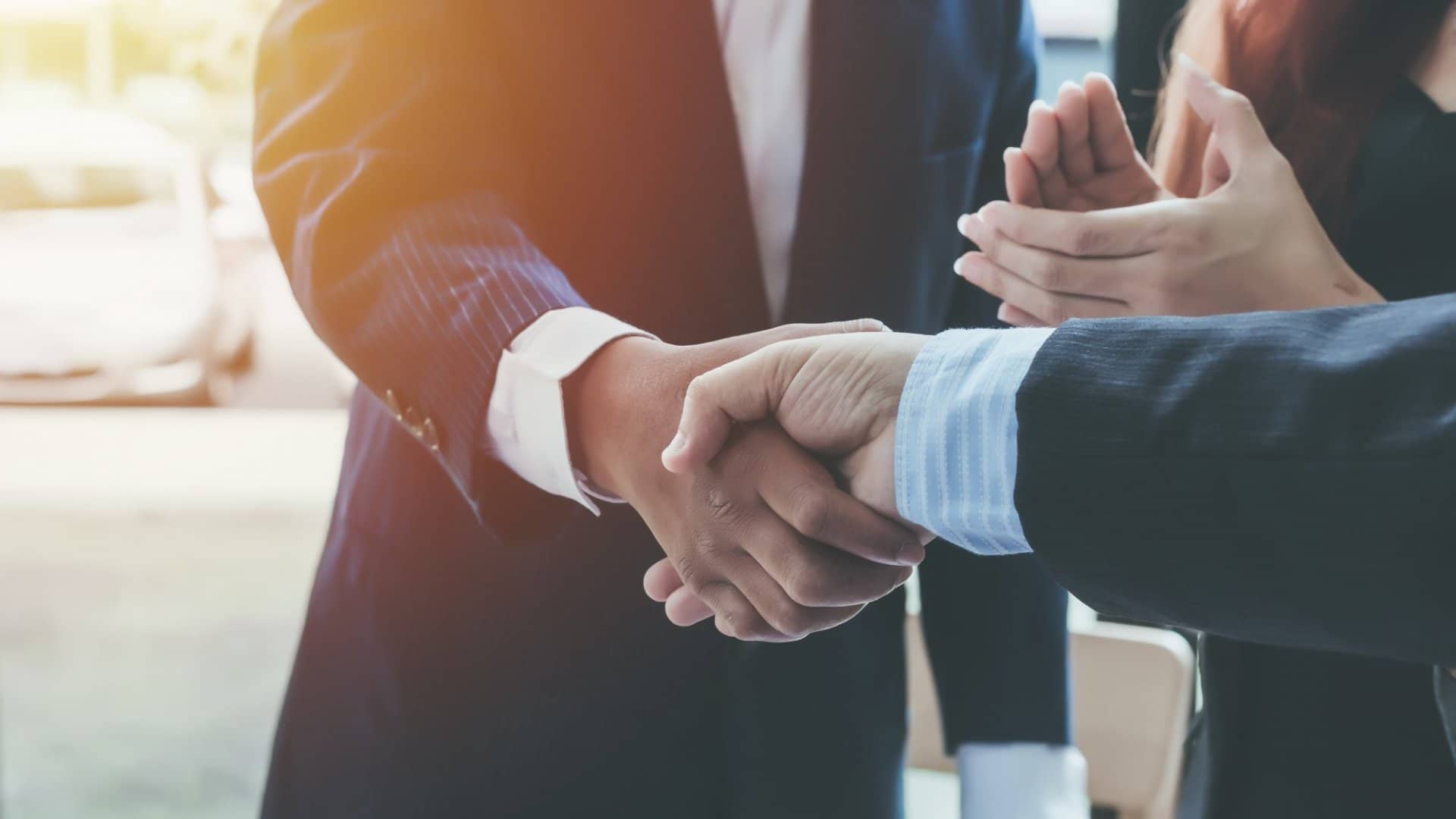 Close-up of a business handshake with clapping hands in the background, indicating agreement or congratulations.